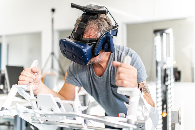Photo of an employee with VR glasses. He is lying on the Icaros fitness device and holding on to hand attachments.
