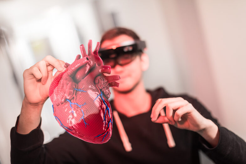 Foto von einem Studierenden mit VR-Brille, der die Hände zur Steuerung in die Höhe hält. Vor ihm sieht man, was er durch die Brille sieht: ein 3D-Herz-Hollogramm. __ Student wears VR glasses and holds hands up to control. In front of him you can see what he sees through the glasses: a 3D heart hollogram.