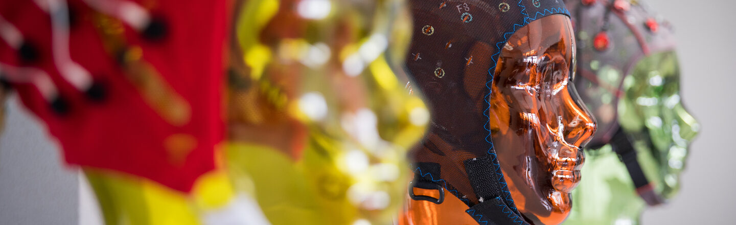 Foto von verschiedenen EEG-Masken mit Ständer in Form von Köpfen auf einem Regal. Im Fokus ist ein orangener Glaskopf in der Mitte. __ Various EEG masks with stands in the shape of a face are lined up on a shelf. The focus is on the orange glass head in the middle.