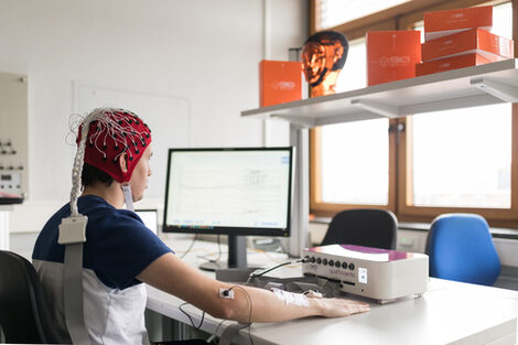 Photo of a test person with EEG cap and neuroreceptor on elbow in front of a measurement screen.