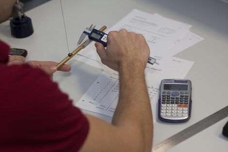 Photo of a person measuring the diameter of a pipe with calipers. On the table are pieces of paper with notes, a calculator and a ruler.