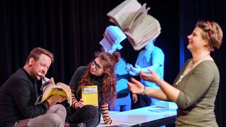Photo of actors performing a scene in the theater lab while juggling books.