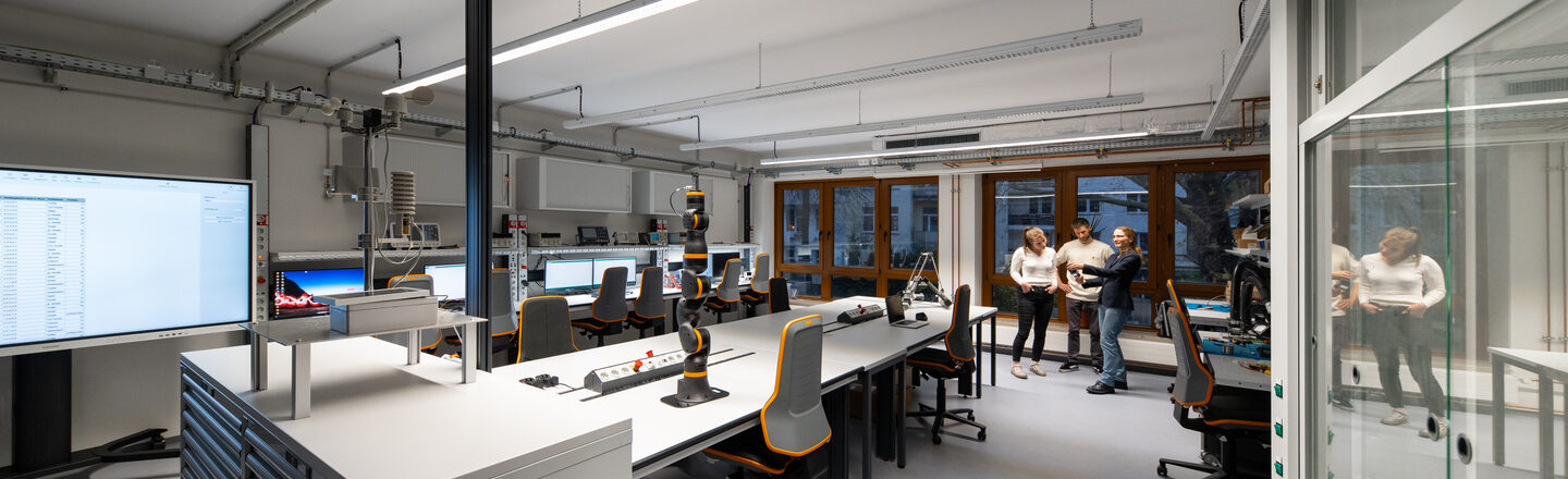 You can see a modern laboratory with several workstations equipped with computers, monitors and technical equipment. In the middle of the room are workbenches with ergonomic chairs, power strips and a robotic arm. In the background are three people having a conversation.