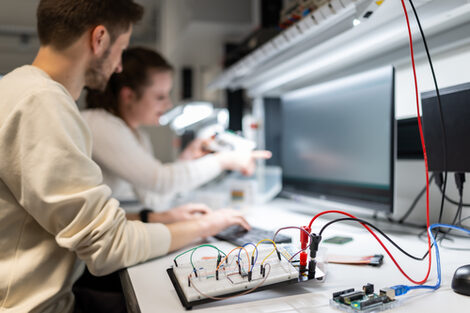 Two people are working on an electronic circuit in a laboratory. In the foreground there is a breadboard with cables and components on the table. The workstation is equipped with measuring devices, tools and lighting.