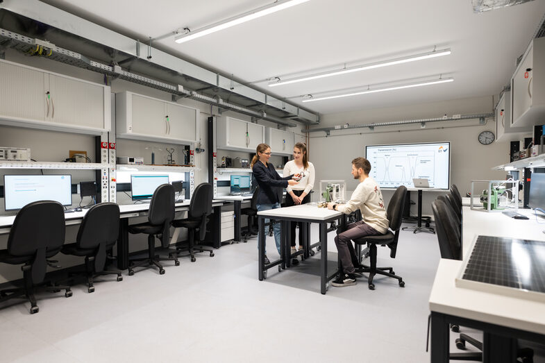 You can see a modern laboratory with several workstations equipped with computers, monitors and technical equipment. In the middle of the room you can see a person sitting at a table and two people standing in front of her, talking to each other.