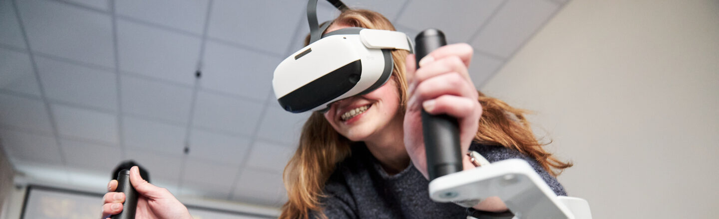 Photo of a person on the Icaros device. She is wearing VR goggles and holding on to the handles.