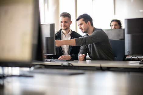 Foto eines Dozenten neben einem Studenten am PC. Der Dozent zeigt dem Studenten etwas auf dem Bildschirm. Im Hintergrund ein weiterer Student an einem Computer.