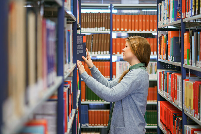 Photo of a female student in a row of books in the library. She is facing the left shelf and is taking out a book.