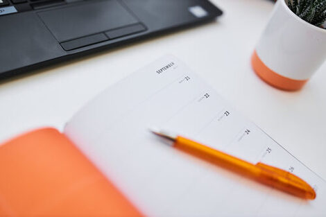 Photo of an open diary and a ballpoint pen lying on a table. Behind them is a laptop and a small flower pot in the cut.