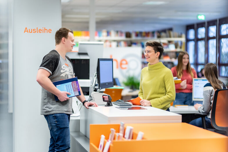 Foto einer Mitarbeiterin hinter der Theke der Bibliothek, sie ist mit dem ihr gegenüber stehenden Studenten, der Bücher in der Hand hat, im Gespräch. Im Hintergrund ist eine weitere Studentin im Gespräch mit einer Mitarbeiterin.