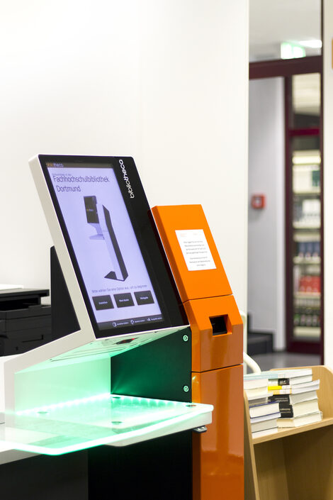 Photo of a self-service check-out machine in the library with a stack of books on a book cart next to it__Photo of a self-service check-out machine in the library with a stack of books on a book cart next to it