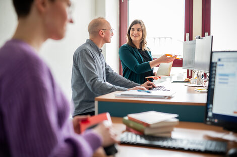 Drei Personen arbeiten in Bibliothek. Zwei sitzen gemeinsam vor einem PC und unterhalten sich. Die dritte Person scannt ein Buch an einem anderen PC.