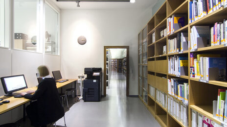 Foto eines Raums in der Bibliothek, in dem sich ein Bücherregal, ein Multifunktionsgerät zum Kopieren, Drucken und Scannen sowie mehrere Computer-Arbeitsplätze befinden__Photo of a room in the library featuring a book shelf, a multi-purpose device for photocopying, printing and scanning, and several computer desks