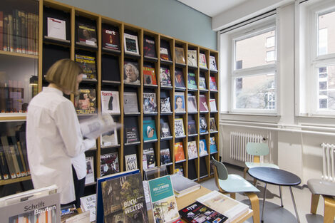 Foto einer Studierenden vor dem Zeitschriftenregal in der Bibliothek. In dem Raum befinden sich auch eine Sitzecke, ein Tisch mit Büchern und eine Vitrine mit historischen Büchern__Photo of a female student standing in front of a library journal shelf. The room also features chairs, a table with books and a display cabinet with historic books