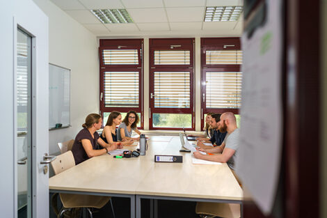 Photo in a room. Six students at a group table are exchanging ideas. Working materials are lying on the table __View into a room. Six students at a group table exchange ideas. Working materials are on the table.