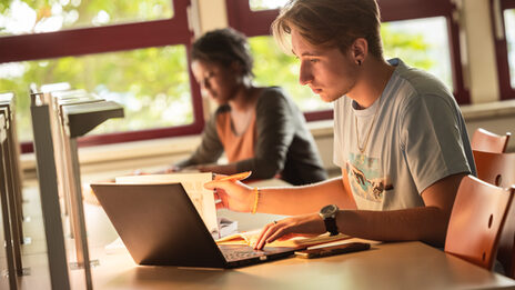 Photo of two students studying with a laptop and books in the library's reading room.