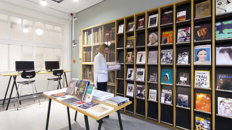Photo of a female student standing in front of a library journal shelf. The room also features a table with books, a display cabinet with historic books and two computer desks__Photo of a female student standing in front of a library journal shelf. The room also features a table with books, a display cabinet with historic books and two computer desks