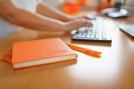 Foto von einem FH-Notizbuch und FH-Kugelschreiber, die auf einem Schreibtisch liegen. Im Hintergrund tippt eine Frau auf der Tastatur. __ FH notebook and FH ballpoint pen are on the desk, a woman is typing on the keyboard in the background.