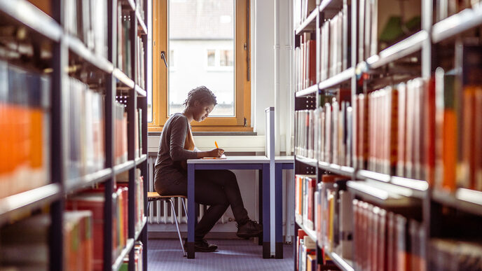 Photo of a young woman sitting at a table in the library between bookshelves and taking notes.