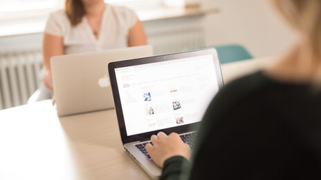 Foto einer jungen Frau am Laptop, ihr gegenüber am Tisch sitzt eine weitere Frau mit Laptop. __ Young woman at the laptop, sitting across from her at the table, another woman with a laptop.