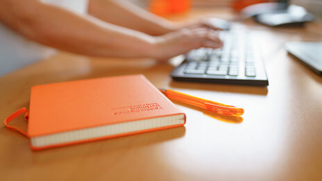 Foto von einem FH-Notizbuch und FH-Kugelschreiber, die auf einem Schreibtisch liegen. Im Hintergrund tippt eine Frau auf der Tastatur. __ FH notebook and FH ballpoint pen are on the desk, a woman is typing on the keyboard in the background.