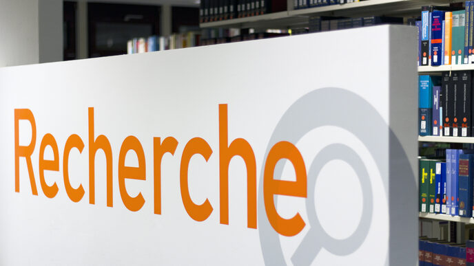 Photo of a sign reading "Recherche" (German for "research") with a library book shelf in the background__Photo of a sign reading "Recherche" (German for "research") with a library book shelf in the background
