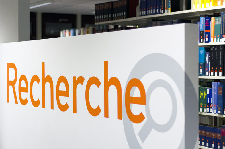 Photo of a sign reading "Recherche" (German for "research") with a library book shelf in the background__Photo of a sign reading "Recherche" (German for "research") with a library book shelf in the background