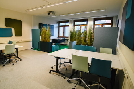Foto der BibLounge am Standort Sonnenstraße mit Tischen, Stühlen, einem Kicker und Kunstpflanzen.__Photo of the BibLounge at campus Sonnenstraße with tables, chairs, a football table and artificial plants.