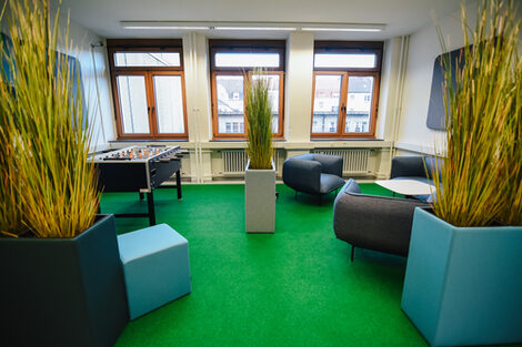 Foto der BibLounge am Standort Sonnenstraße mit einem Kicker, Sesseln, einem Sofa und Kunstpflanzen__Photo of the BibLounge at campus Sonnenstraße with a football table, armchairs, a sofa and artificial plants.