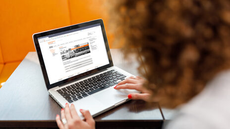 Photo of a young woman at the laptop, in the background there are orange colored chairs. __ Young woman at the laptop, in the background there are orange colored chairs.