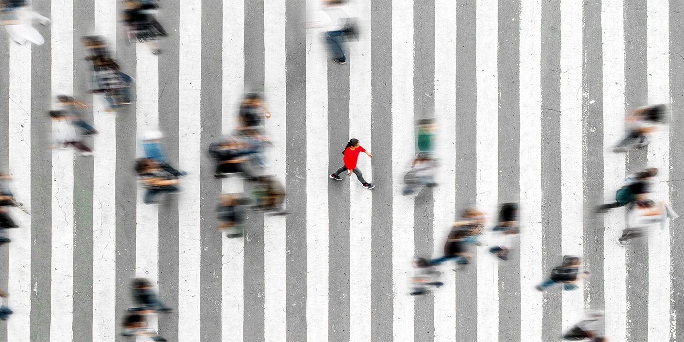 View from above of a large crosswalk on a road. Many people are walking across it. They are blurred. A person in a red sweater is in sharp focus in the middle.