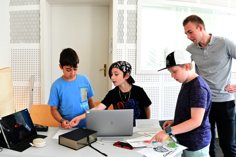 Drei Schüler stehen um einen Tisch mit Notebook und verschiedenen Utensilien, um das Rätsel um die „Digitale Rettung“ zu lösen. Ein Tablet zeigt die verbleibende Zeit von 41:33 an und ein Mitarbeiter der FH gibt einen Hinweis zur Lösung des Rätsels.