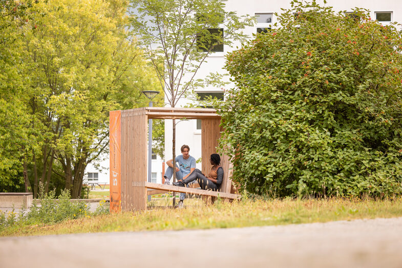 Foto von 2 Studierenden, die in einem Holzwürfel, der mit Sitzgelegenheiten ausgestattet ist, sitzen und sich unterhalten. In der Mitte des Würfels, ist ein Baumgepflanzt.