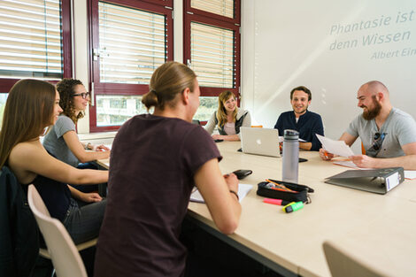 Foto von sechs Studierenden an einem Gruppentisch, die sich austasuchen. Auf dem Tisch liegen Arbeitsmaterialien.__Six students at a group table exchange ideas. Working materials are on the table.