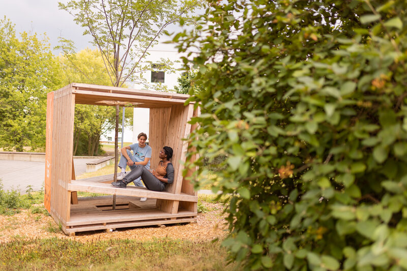 Foto von 2 Studierenden, die in einem Holzwürfel, der mit Sitzgelegenheiten ausgestattet ist, sitzen und sich unterhalten. In der Mitte des Würfels, ist ein Baumgepflanzt.