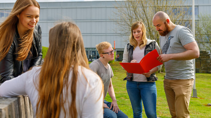 Photo of students on campus looking at a folder together and exchanging ideas. In the foreground two other students.__One female and tweo male students sit and stand on the campus, one student holds a folder in his hand and they exchange ideas. In the foreground two other students.