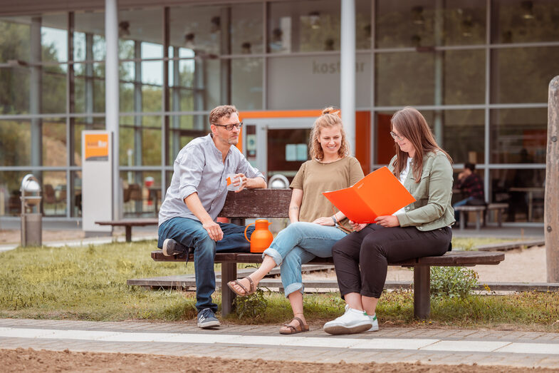 Foto von 3 Personen, die draußen vor der kostBar auf einer Holzbank sitzen und sich besprechen.