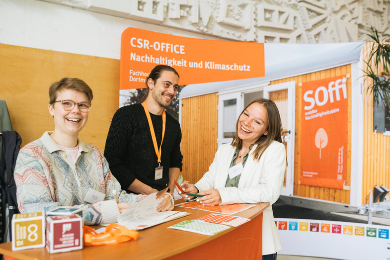 Three receptionists stand behind the counter and laugh amiably into the camera