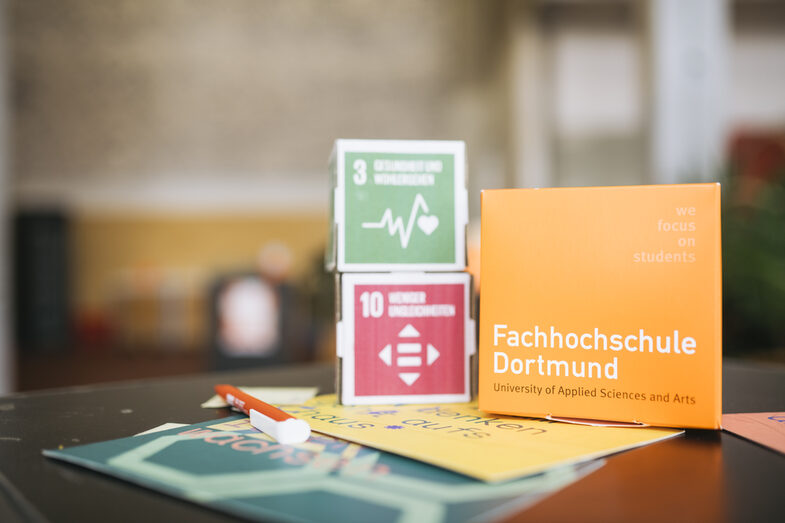 The picture shows a close-up of a FH cardboard cube and two cubes with the sustainability targets 3 and 10 on a table. Brochures and flyers lie in front of the cardboard cubes