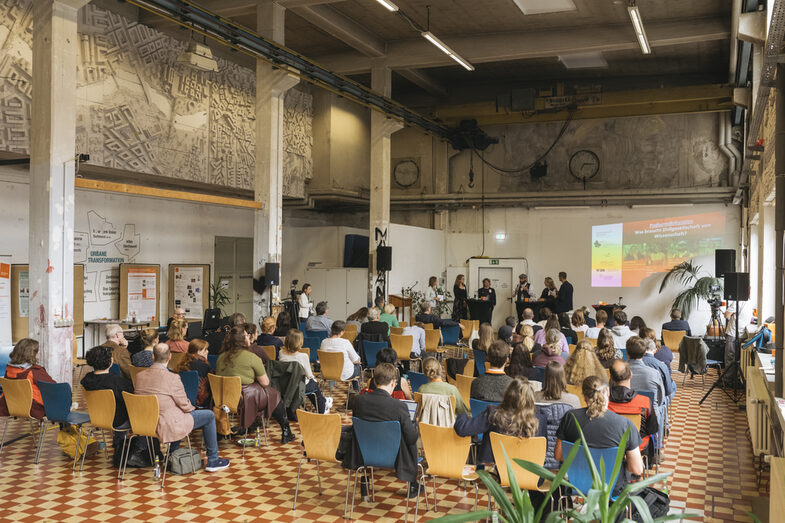 The picture shows a long shot of the Werkhalle venue. Many people are photographed from behind in rows of chairs listening to a lecture. The room is high, large and bright