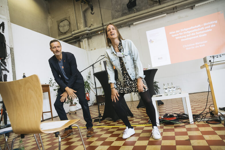 The picture shows two presenters performing synchronized sporting movements. A screen can be seen in the background. The people appear to be looking into an audience.