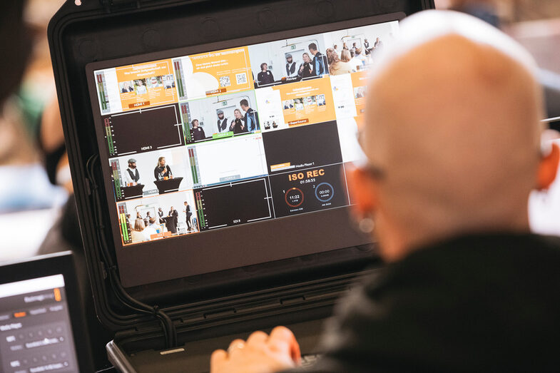 In the picture, a person is sitting with their back to the camera and looking at a screen with several small tiles. It looks like small Viedo recordings