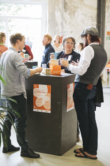 Three people are standing at a dark, square bar table in a bright room, chatting animatedly