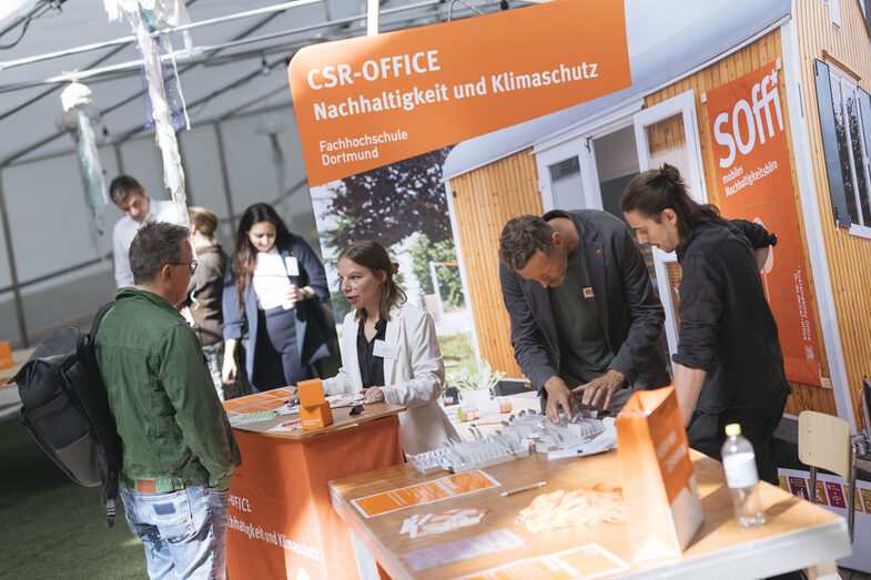 The picture shows a reception desk. You can see a receptionist in a friendly conversation with another person standing with their back to the camera. There is an advertising wall in the background. It says "Fachhochschule Dortmund" on it