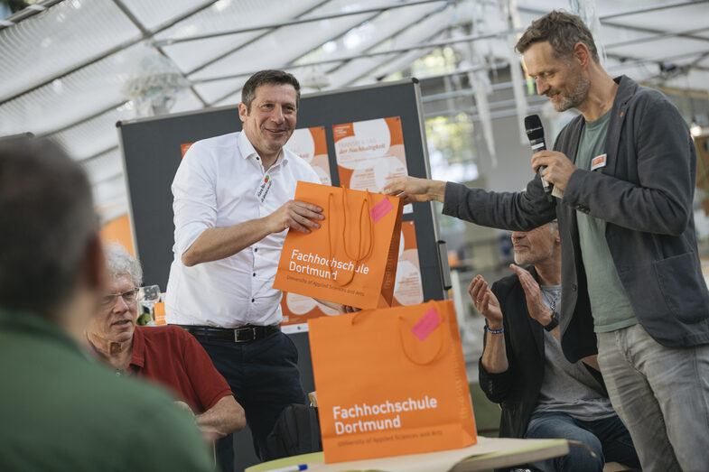 The picture shows two people. One person is handing the other an orange bag with the words Fachhochschule Dortmund on it. The person giving the bag is holding a microphone and speaking to an audience. Blurred outlines of people sitting can be seen at the front left.