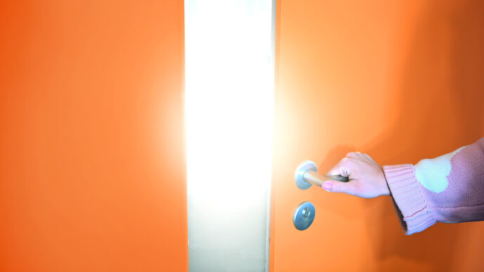A hand grips the handle of a slightly open orange door that reveals a bright light.