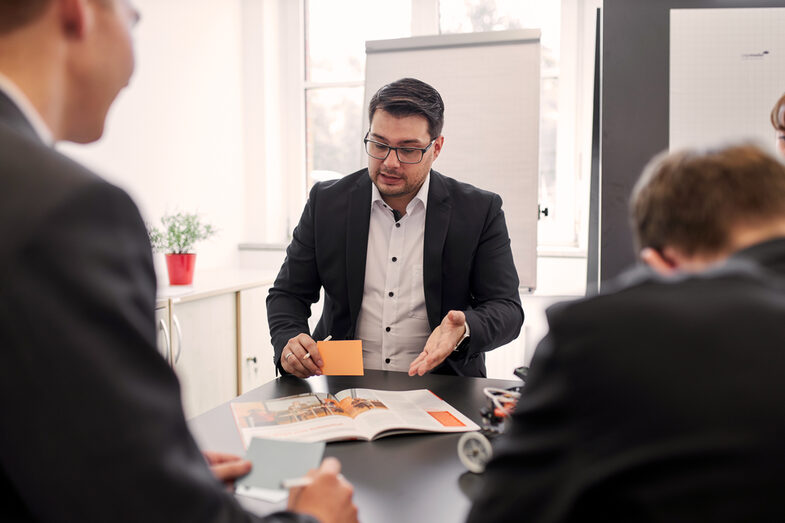 In this photo, four people have gathered at a table. You can see that two of the people are holding index cards and a ballpoint pen. One person with the index cards is also pointing to a magazine and wants to explain something about it.