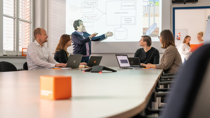 Foto einer Besprechungssituation mit 5 Personen. 4 sitzen an Laptops und eine Frau präsentiert im Stehen. 2 Frauen sind im Hintergrund in einem weiteren Raum erkennbar.