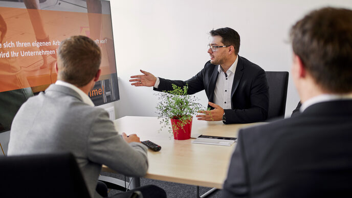 Four people are sitting at a table in a conference room. At the end of the table is a large television with a conference camera attached to it. One of the people is talking and also has his arm outstretched to point at something during the presentation. The others are listening to him.