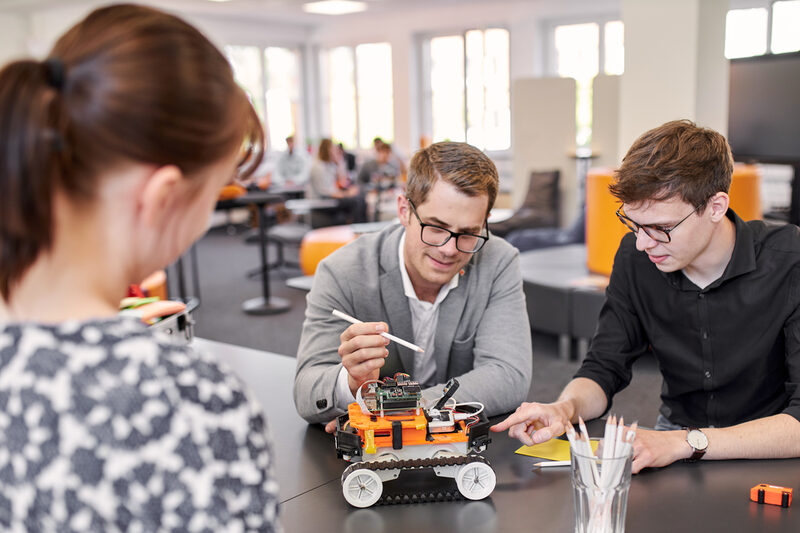 Two people are sitting at a table on which the 4-wheeled robot is standing. Both are leaning slightly forward and both are pointing towards the robot. One is pointing at something with a pencil and the other with a finger. You can also see a person standing on the opposite side of the table, attentively watching the other two people.